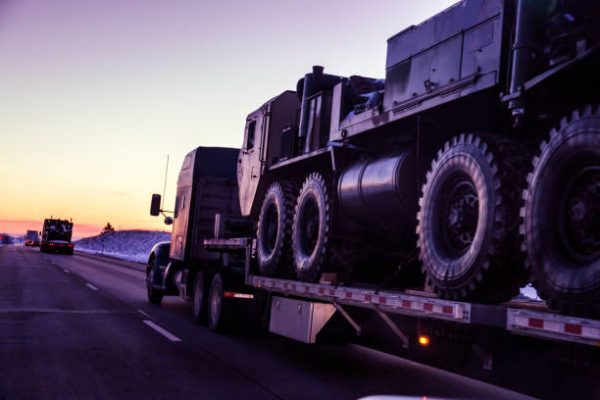 Heading west into a dusky winter sunset, a convoy of semi tractor trailer flatbed trucks loaded with heavy, oversized United States Army tactical armored military land vehicles is hauling the freight along a remote stretch of expressway through the western USA state of Utah. NOTE: Vehicle details have been altered - with various subtractions and/or additions - to make them generic.