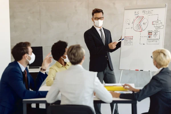 male-entrepreneur-with-face-mask-giving-business-presentation-his-coworker-office